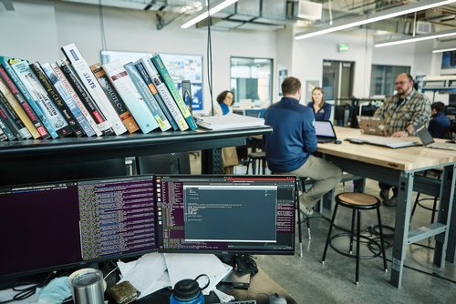 picture of a computer station and monitor in the foreground with four students sitting around a table in a lab in the background 