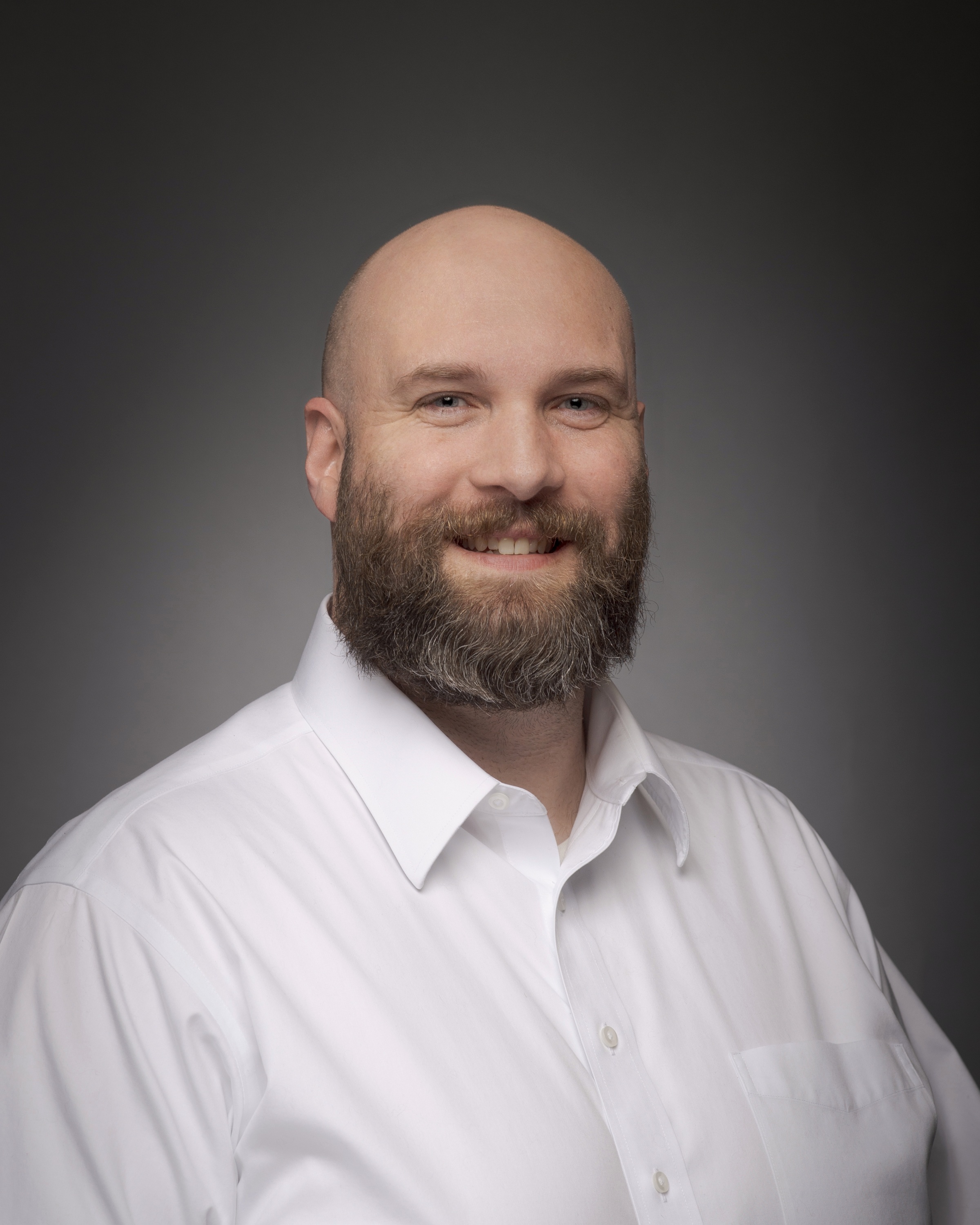 headshot of a bald man with a mustache and beard wearing a white button down shirt in front of a gray background 