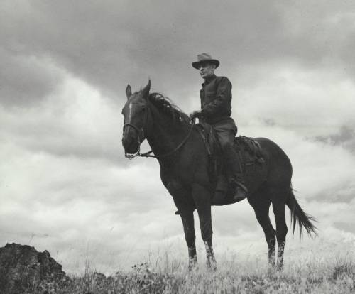 Elers Koch wears dark clothing and sits on a dark horse. The sky is dark in the background.