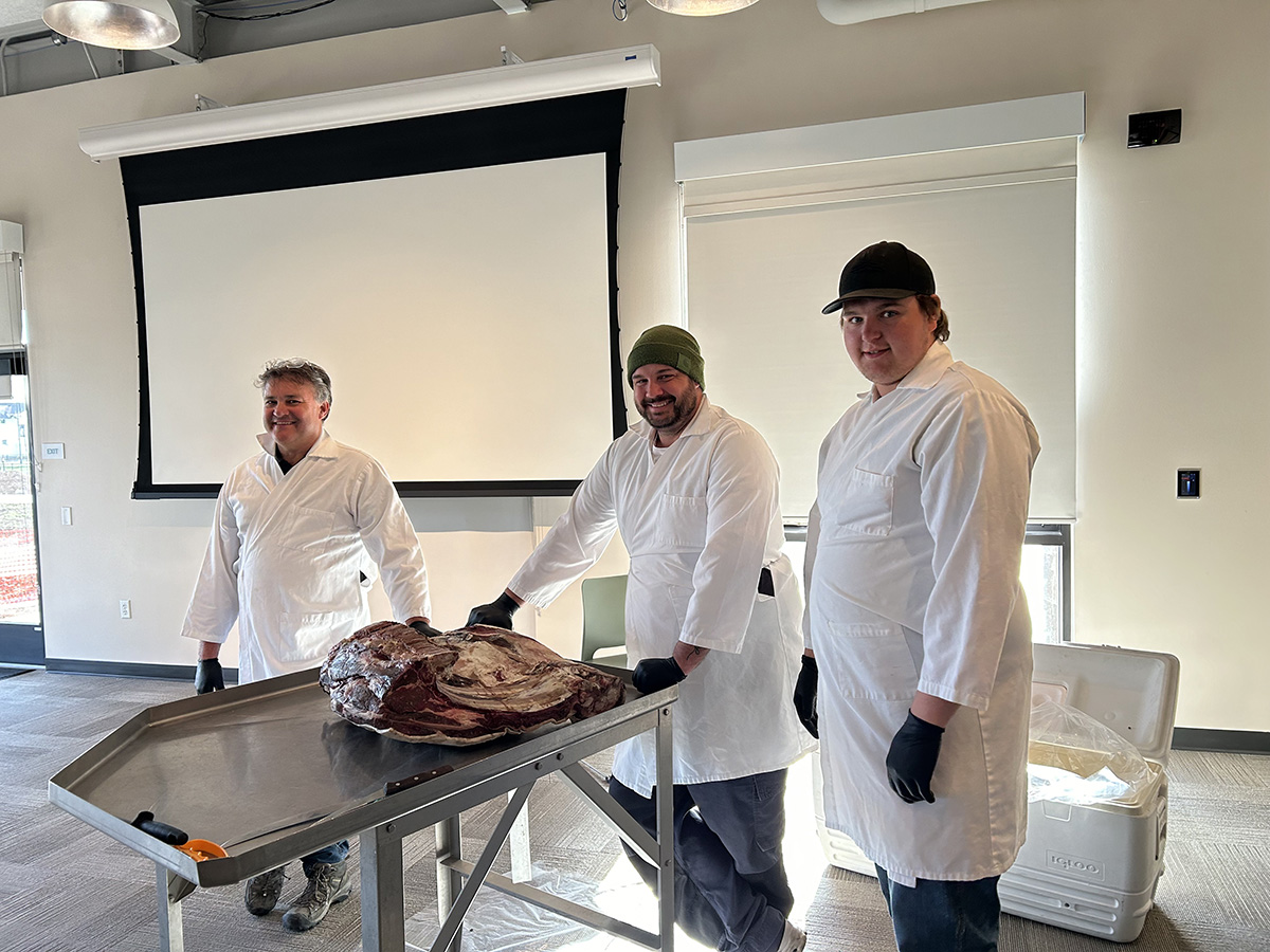 Presenters at a butcher table show an example slab of meat to cut for cooking.