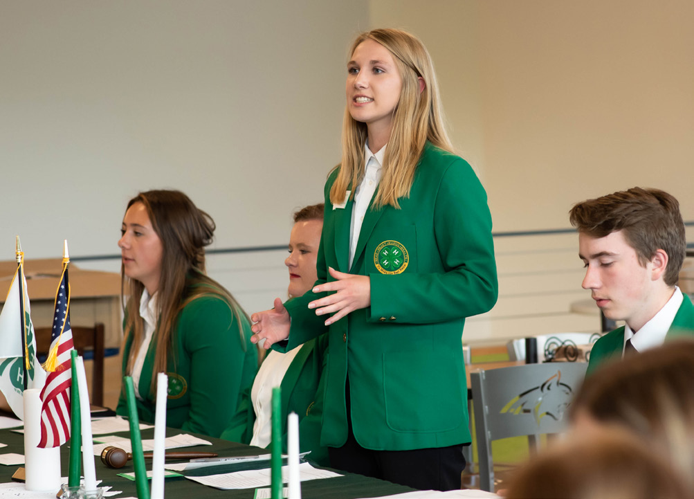 A 4-H leader stands and is speaking to a group of people. She is wearing a green 4-H blazer and is surrounded by other 4-H members in the same blazer.